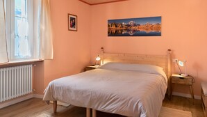A bedroom with a wooden bed, two bedside tables with lamps, a window with curtains, a radiator, and a framed picture on the wall.