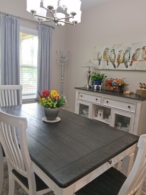 A dining area with a dark wooden table, white chairs, a chandelier, a flower vase, a cabinet with dishes, and a bird painting on the wall.