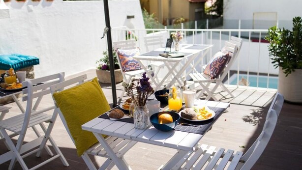 Un área de comedor exterior blanca con una mesa preparada para el desayuno, rodeada de sillas blancas y una piscina.