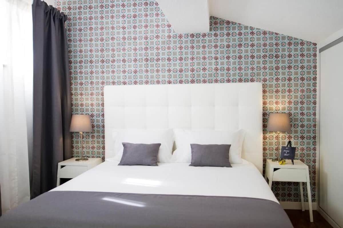 Habitación de hotel con una cama blanca, dos almohadas, una mesita de noche blanca, una lámpara y papel tapiz estampado.