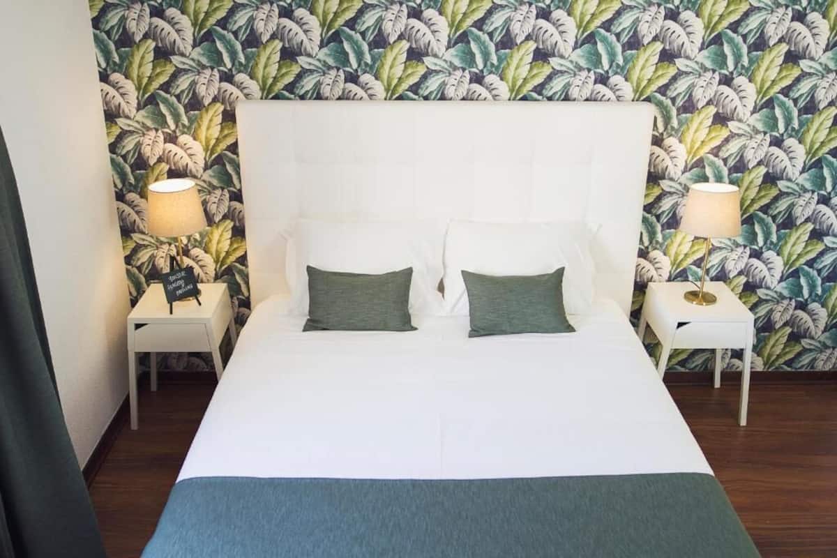 Una cama con cabecera blanca, dos almohadas y una manta verde. Se observan dos mesitas de noche con lámparas y un fondo de papel pintado con motivos florales.