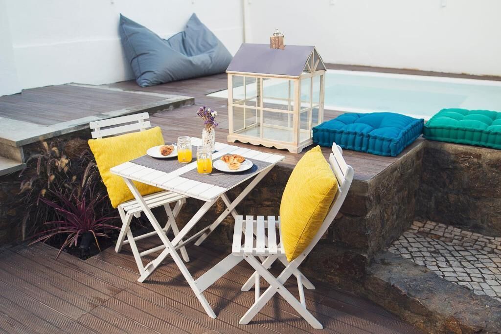 Un área de comedor al aire libre con mesa, sillas, cojines, un farol y una piscina de fondo.