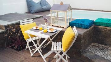Un área de comedor al aire libre con mesa, sillas, cojines, un farol y una piscina de fondo.