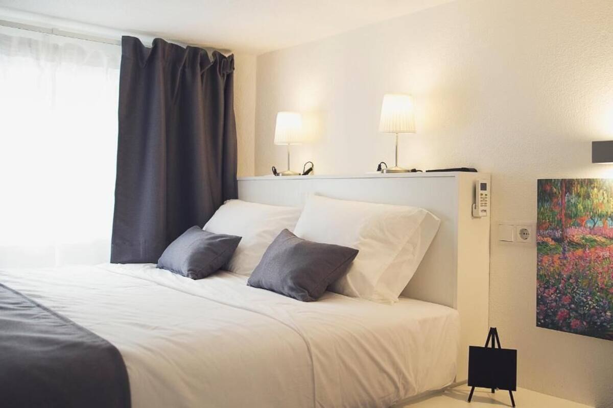 Una habitación de hotel con cama, dos lámparas de noche, un cuadro y una mesita.