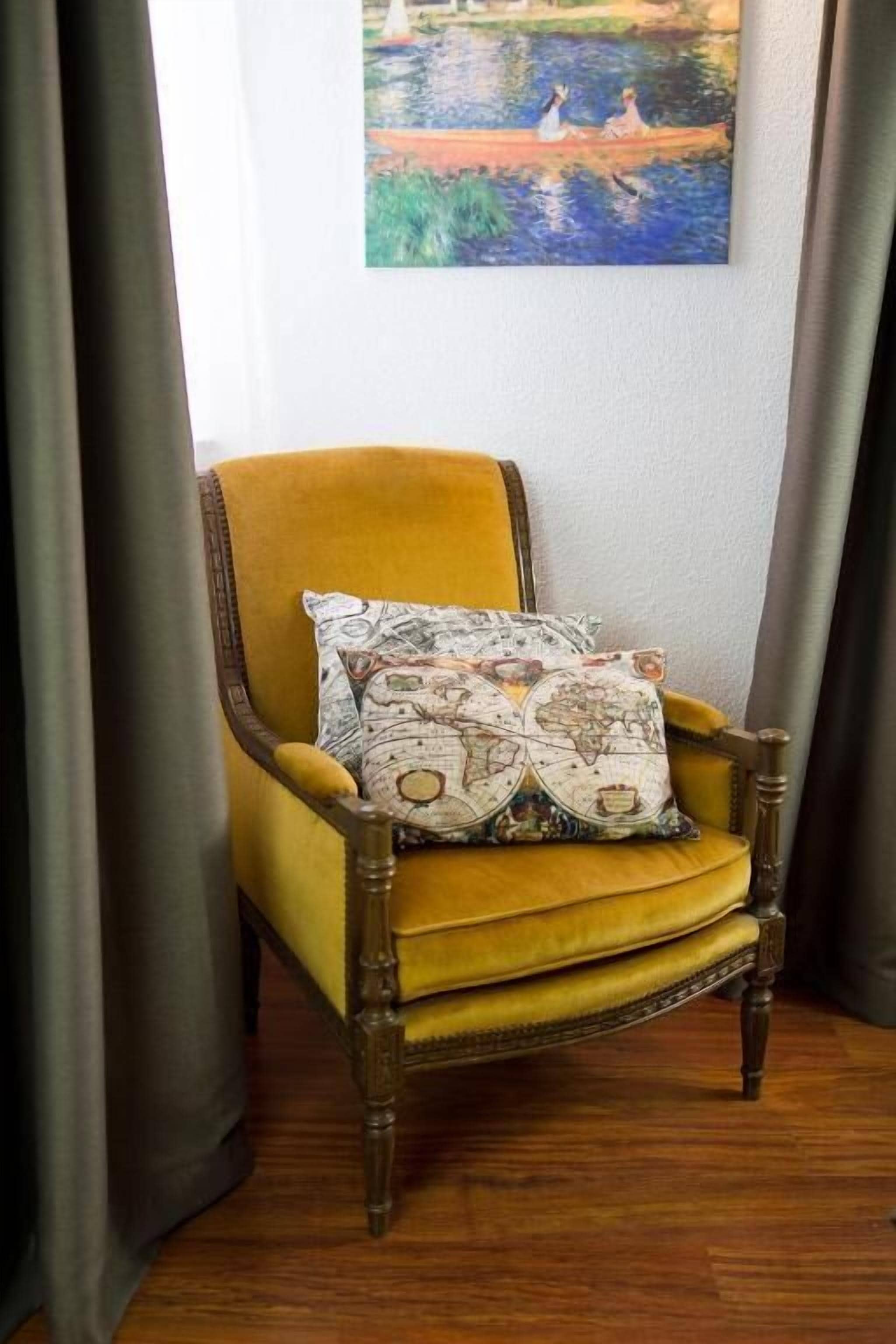 A yellow armchair with a world map pillow, positioned in a room with a painting of a pond and figures on the wall.