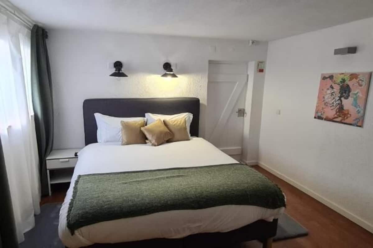 Una habitación de hotel con una cama, una mesita de noche, una lámpara, un cuadro en la pared y una ventana con cortinas.