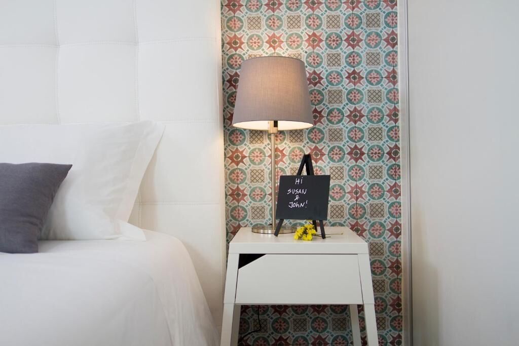 Una cama bien hecha con una almohada, una mesita de noche con una lámpara y una pared decorativa con un diseño geométrico.