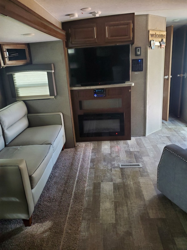 Interior compacto de autocaravana con sofá, televisor de pantalla plana, armarios de madera y un cartel de bienvenida.