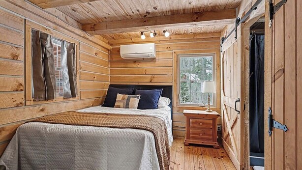 Habitación de cabaña de madera con cama, mesita de noche, ventana y unidad de aire acondicionado.