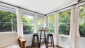 Une pièce avec un plancher en bois, un canapé beige, deux tabourets noirs, une petite table et de grandes fenêtres avec des rideaux.