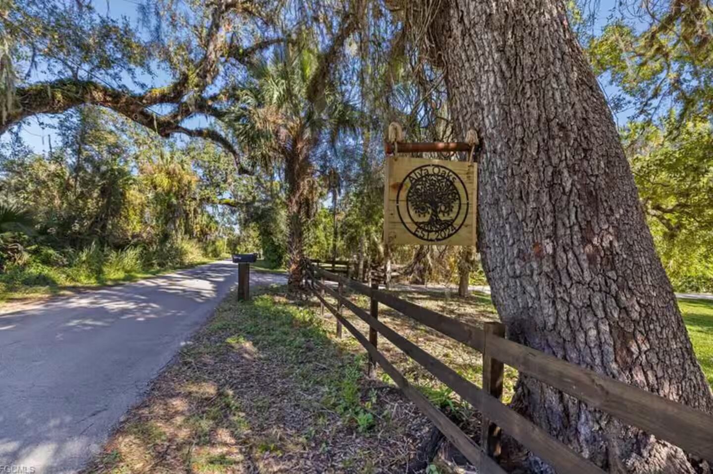 A wooden sign with a tree logo, a mailbox, and a fence along a roadside.