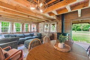 A log cabin interior with a wooden ceiling, a round dining table, and a large window overlooking a green landscape.