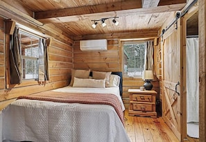 A log cabin bedroom with a bed, bedside table, lamp, and an air conditioning unit.
