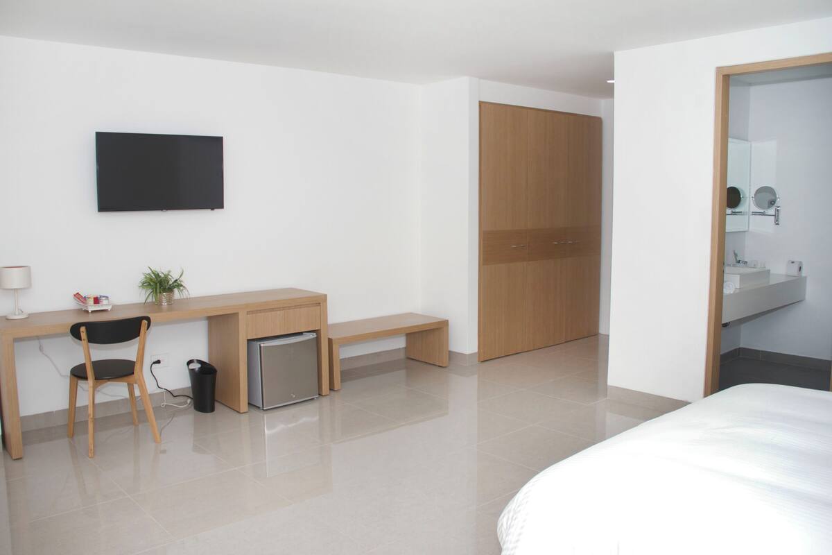 Una habitación de hotel moderna con cama, escritorio con silla, un pequeño cubo de basura, televisor y armario de madera.