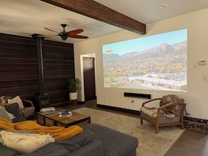 A living room with a fireplace, a ceiling fan, a projector screen displaying a landscape, and a couch with a throw blanket. - 3BR Farmhouse in Durango with Fitness Room, Sauna, Bidets, Theater Entertainment (Durango)