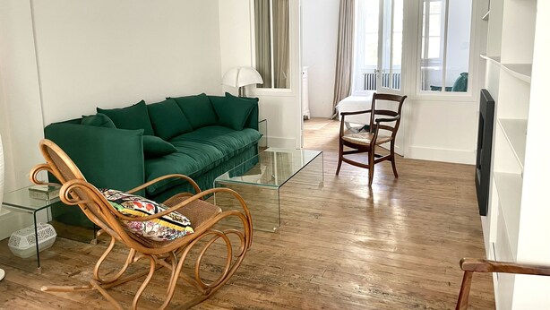 Un salón con un sofá verde, un sillón de mimbre, una mesa de centro de vidrio y suelo de madera.