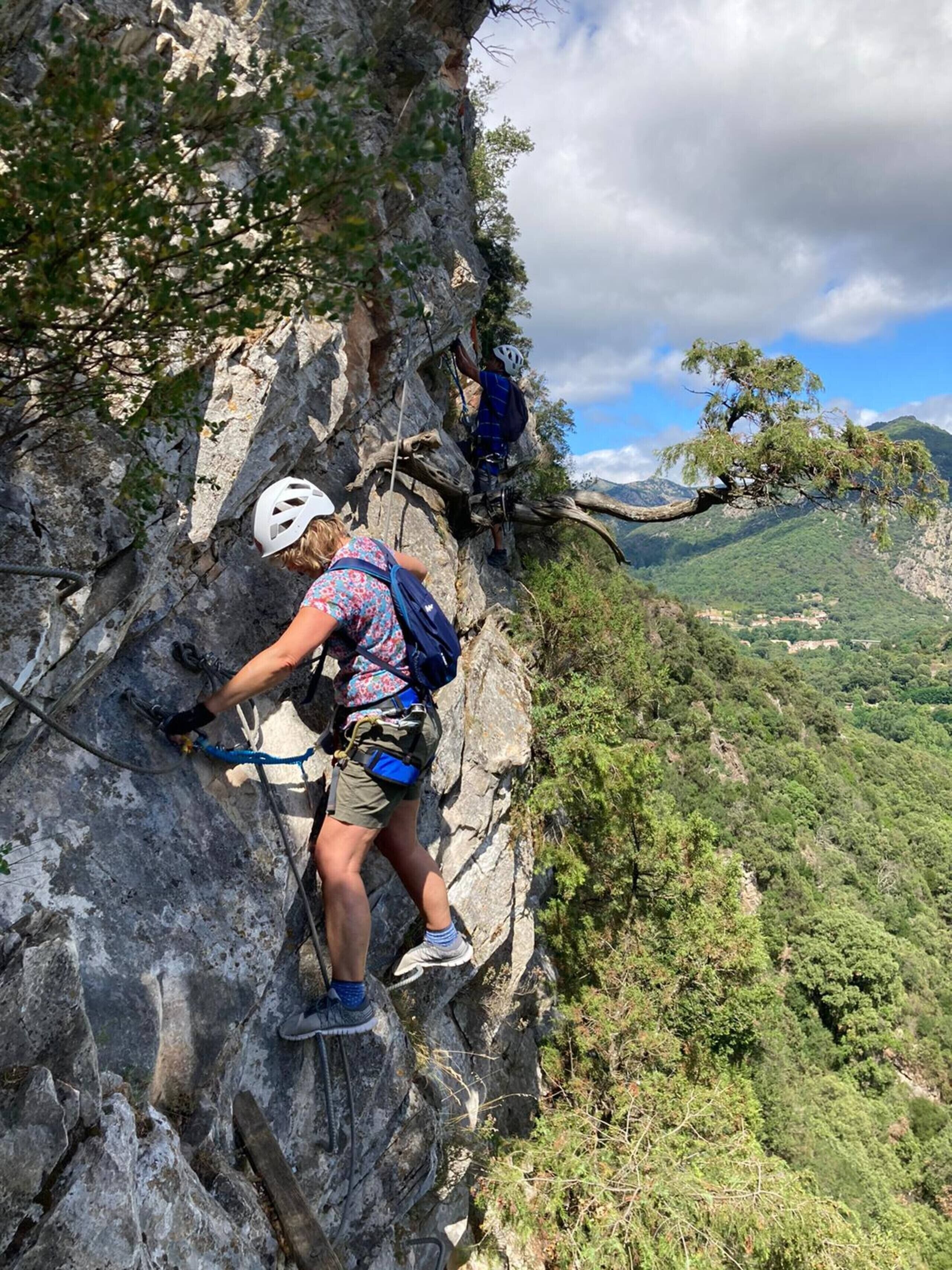 Eine Person erklimmt einen steinigen Bergpfad, im Hintergrund ein Panoramablick auf ein Tal und Bäume.