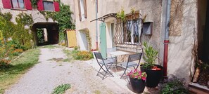 Un patio con una mesa y sillas, un pequeño jardín y un edificio con contraventanas rojas y un balcón.