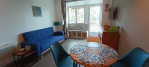 Ein Zimmer mit einem blauen Sofa, einem runden Tisch mit gemusterten Untersetzern, einem kleinen Beistelltisch, einer Lampe und einer Zimmerpflanze.