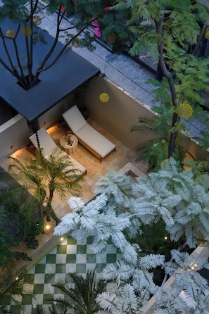 Un espace de détente extérieur, aménagé avec des sièges et une petite table, entouré de palmiers et de fougères, et abrité par une structure ombragée.