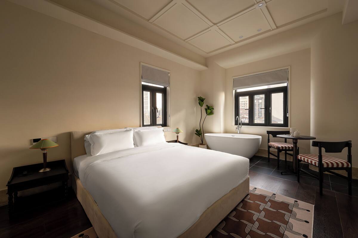 Habitación de hotel con una cama grande, una zona de estar con una silla y una mesa, una bañera y un ventanal con vistas a edificios.