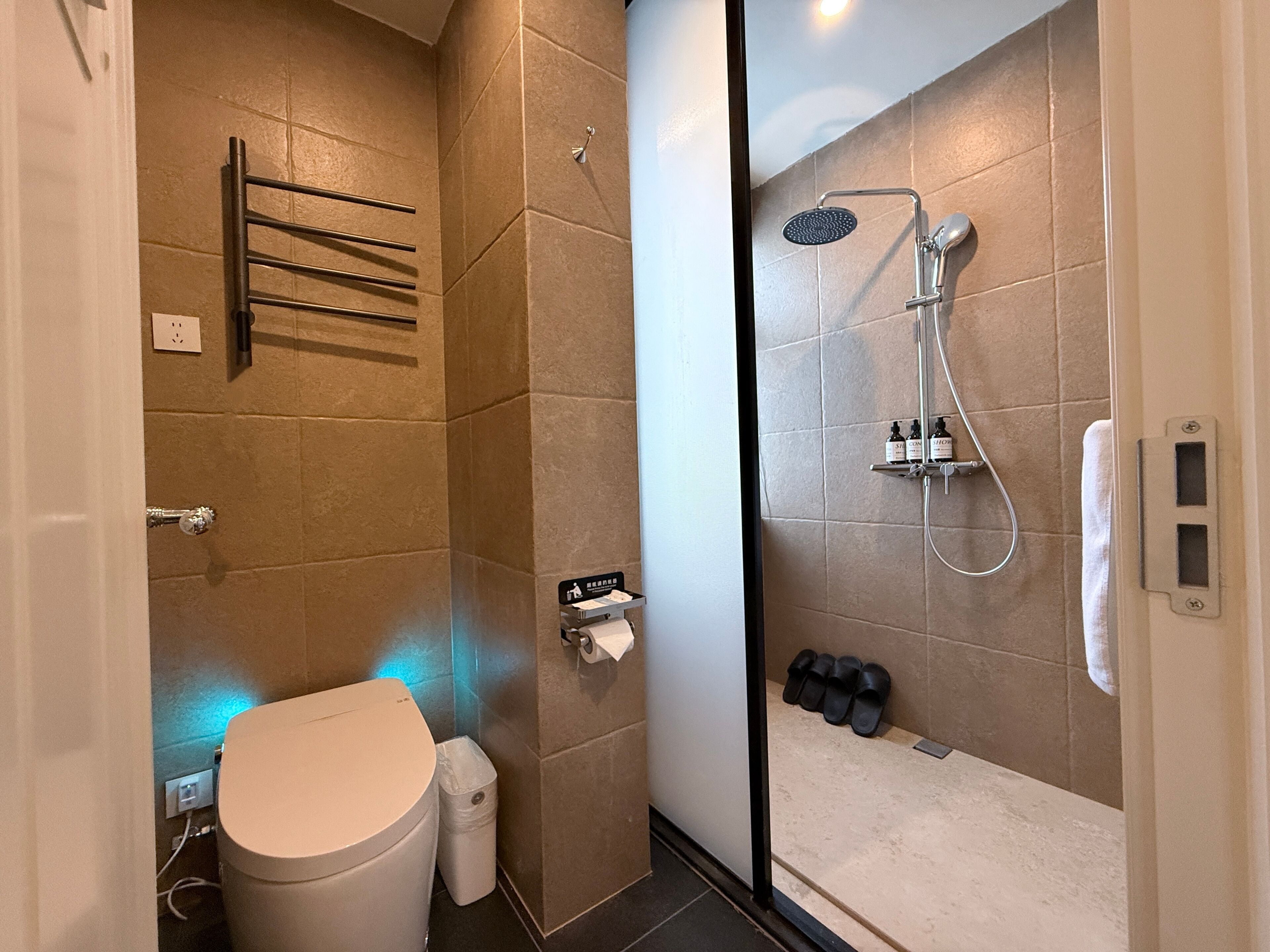 A modern bathroom with a toilet, towel rack, shower, and a pair of slippers.