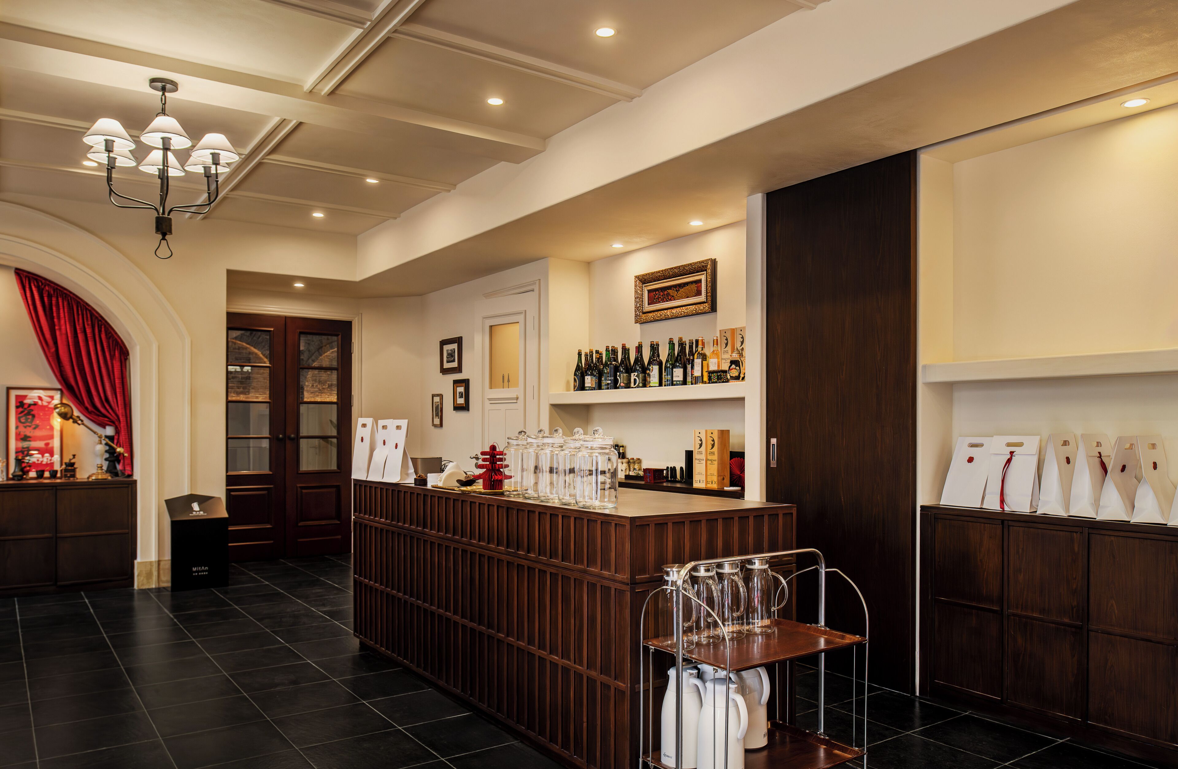 A bar area with a wooden counter, shelves with bottles, a chandelier, and a door with glass panels.
