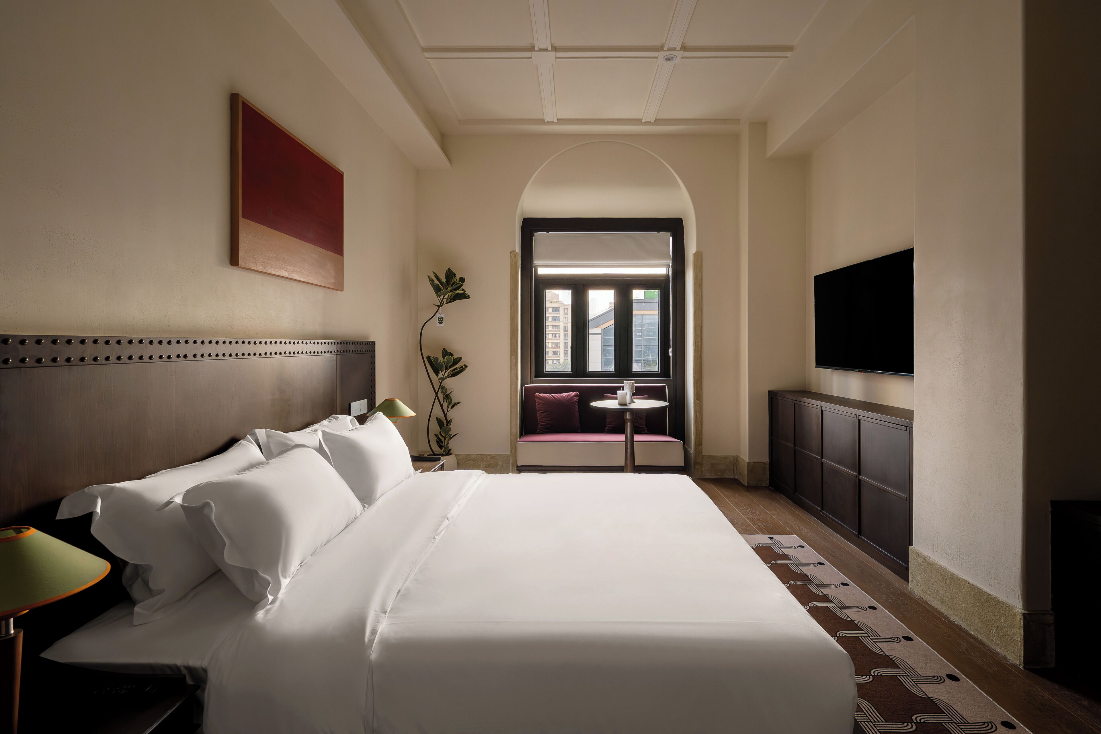 A hotel room with a large bed, a TV, a desk with a chair, a window, and a view of buildings outside.