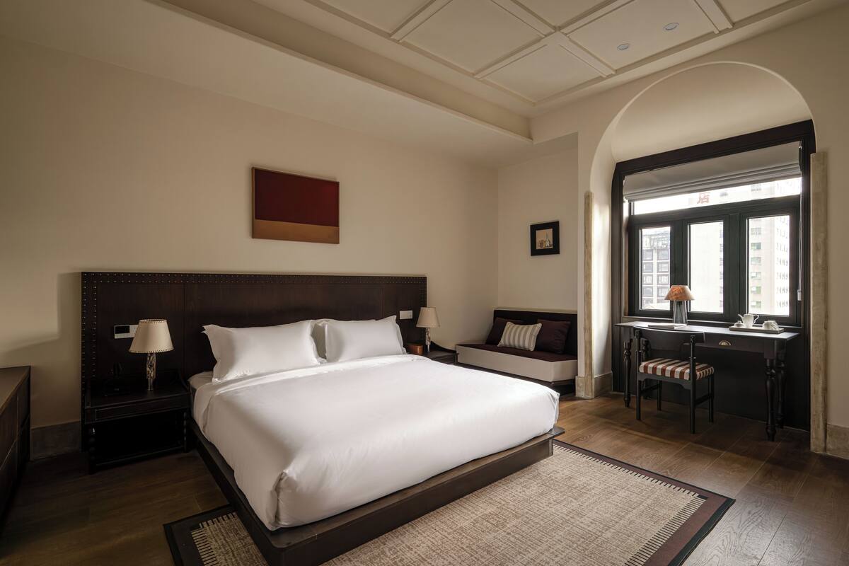 Una habitación de hotel con una cama grande, un escritorio con una silla, una mesita de noche con una lámpara y vistas a edificios a través de una ventana.