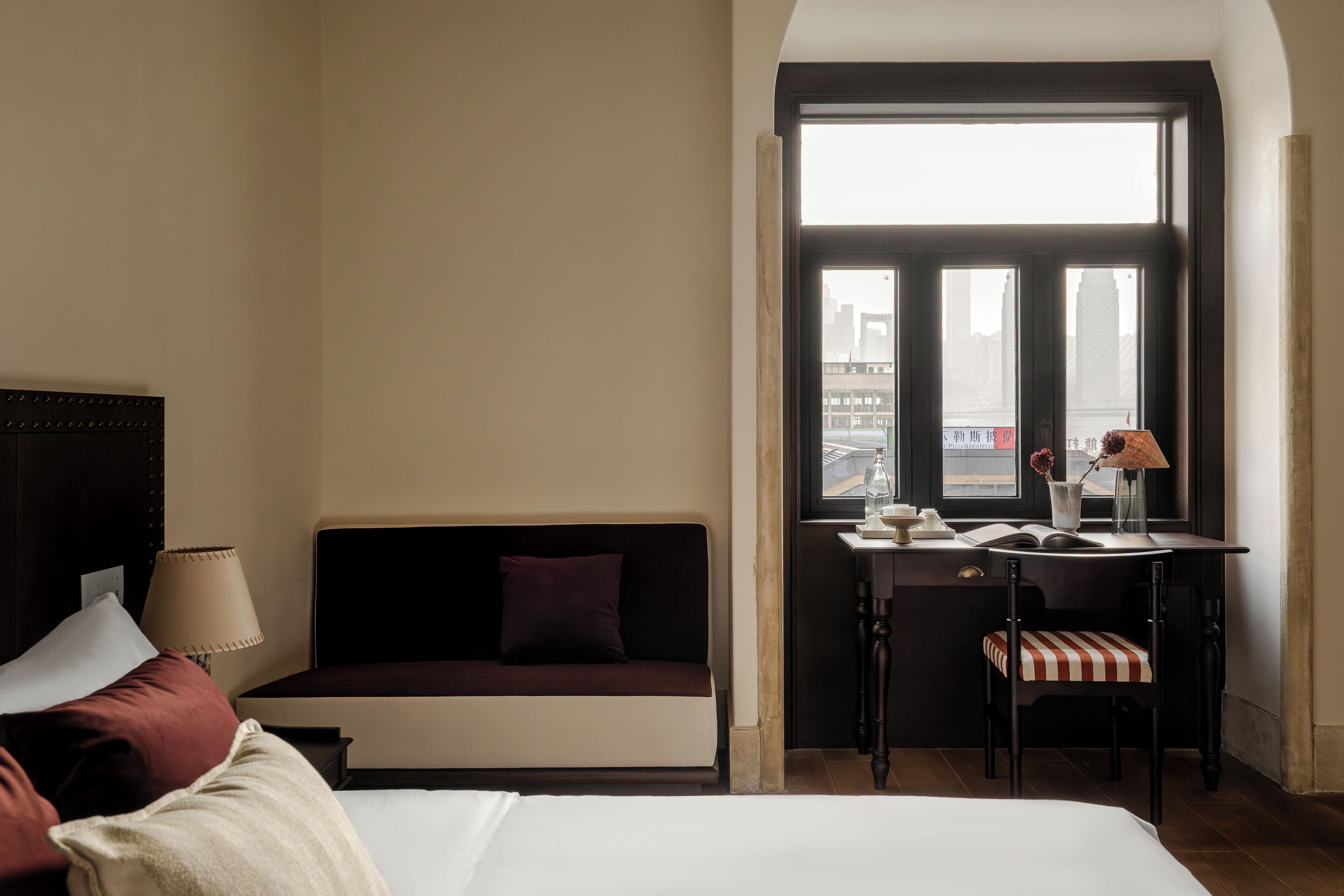 Una habitación de hotel con una cama, un sofá, un escritorio con una silla, una ventana con vistas a edificios y una lámpara.