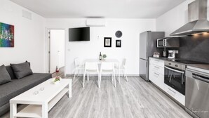 Eine moderne Küche mit weißen Geräten, ein Essbereich mit weißem Tisch und Stühlen und ein graues Sofa in einem hellen Raum mit Flachbildfernseher und Wandbildern.