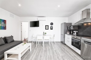 Una cocina moderna con electrodomésticos blancos, un comedor con mesa y sillas blancas, y un sofá gris en una habitación luminosa con televisor de pantalla plana y obras de arte en la pared.