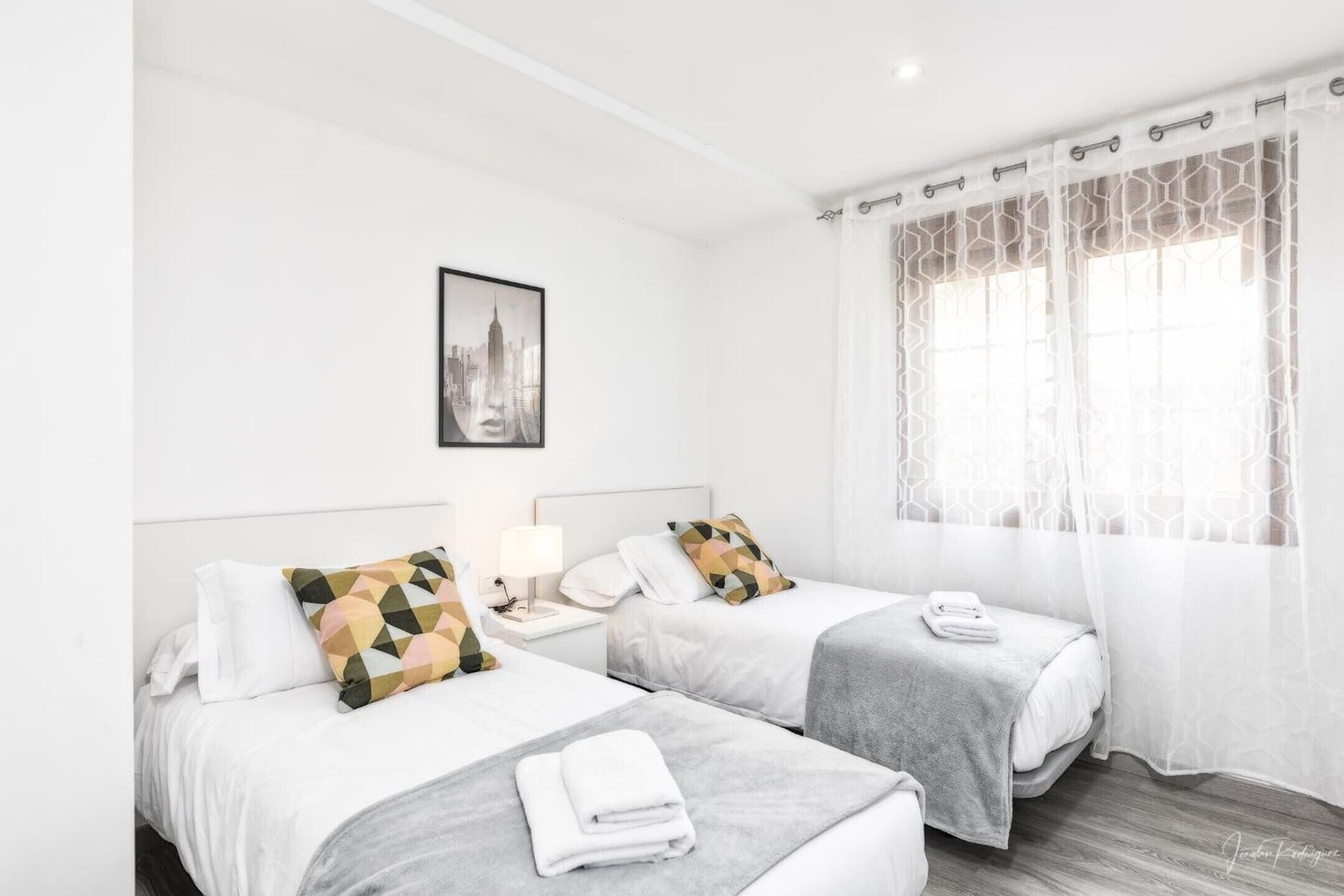 A hotel room with two beds, a nightstand, a lamp, a framed picture on the wall, and a window with sheer curtains.