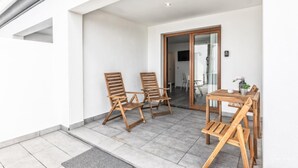 Ein Balkon mit zwei Holzstühlen, einem Tisch und einer Topfpflanze darauf. Eine Tür führt nach innen.