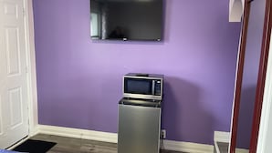 Una habitación con paredes moradas, un televisor de pantalla plana sobre un microondas, una puerta y una cama con colcha azul.
