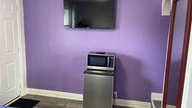 A room with purple walls, a flat-screen TV mounted above a microwave, a door, and a bed with a blue bedspread. - OSHAWA INN MOTEL (Oshawa)