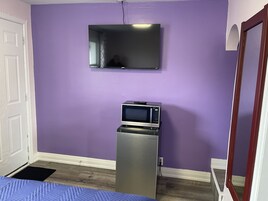 A room with purple walls, a flat-screen TV mounted above a microwave, a door, and a bed with a blue bedspread.