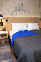 A hotel room with a bed, a nightstand, a lamp, a map on the wall, and a small plant.