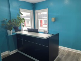 Una habitación con paredes verde azulado, un escritorio negro con una computadora portátil, dos ventanas y una planta en maceta.