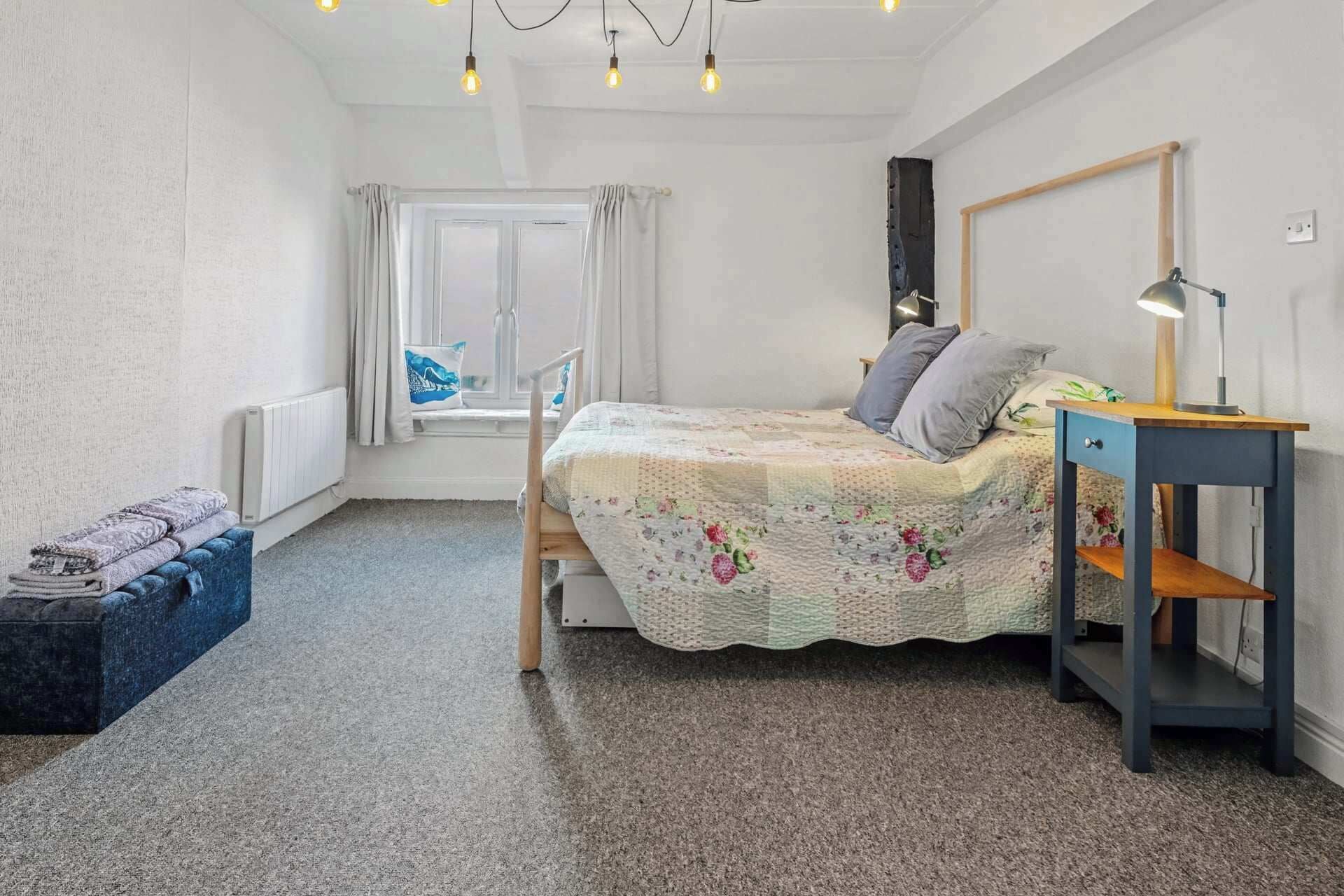 Une chambre à coucher comprenant un lit, une table de chevet, une lampe, une fenêtre avec des rideaux, un radiateur et un petit banc.