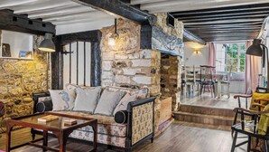 Et rustikt interiør med synlige stenvægge, en træsofa, et spiseområde med stole og en trappe til en etage ovenpå.