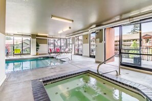 An indoor swimming pool with clear glass walls, a separate hot tub area, and a view of the surrounding buildings.