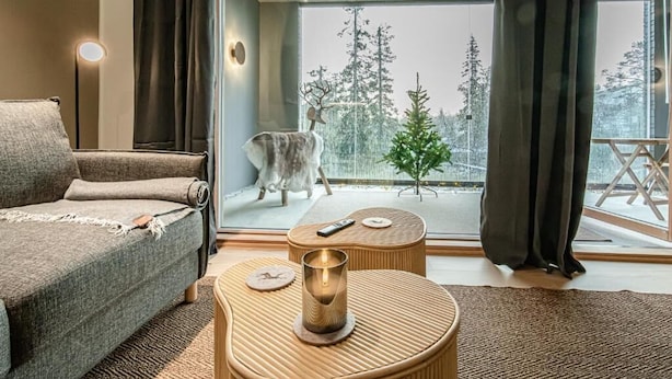 Ein gemütliches Zimmer mit einer Couch, einem kleinen Holztisch mit Kerze und Blick auf einen verschneiten Wald.