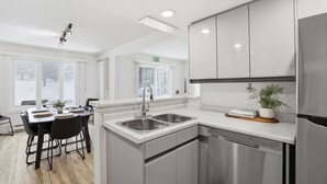 A modern kitchen with stainless steel appliances, a dining area with a table set for four, and a view of the outdoors through large windows. - Modern Escape - Steps to Village + Summer Pool (The Blue Mountains)