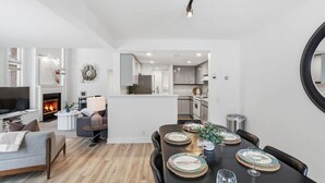A modern living room with a fireplace, a dining area with a table set for four, and a kitchen in the background. - Modern Escape - Steps to Village + Summer Pool (The Blue Mountains)