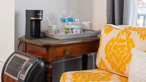 Ein Hotelzimmer mit einem hölzernen Nachttisch, einer schwarzen Kaffeemaschine, zwei Wasserflaschen, einer Ablage mit Toiletteartikeln, einem weißen Kopfkissen und einem gelb gemusterten Kissen.