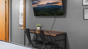 A room with a bed, a wooden desk with two chairs, a TV mounted on the wall, and a small potted plant. - Meadow View | King Suite for 2 | Wears Valley (Sevierville)
