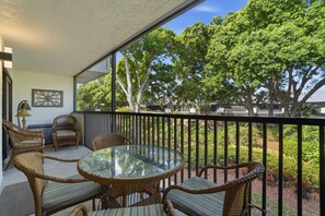 Ein Balkon mit Korbmöbeln, einem Glastisch und Blick auf Bäume und einen Garten.