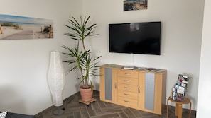 Una habitación con un armario de madera, una planta en maceta, un jarrón grande, un televisor de pantalla plana y un cuadro en la pared.