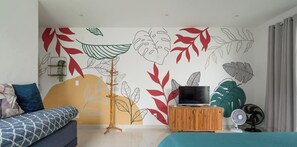 Une chambre à coucher avec un lit, un téléviseur posé sur un meuble en bois, une lampe sur pied et un mur orné de motifs de feuilles.